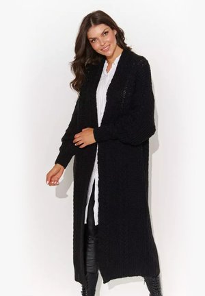 Uśmiechnięta kobieta, ubrana w długi czarny dziergany kardigan na białej koszuli i czarne skórzane spodnie, stoi na białym tle.