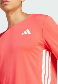 Czerwona sportowa koszulka z okrągłym dekoltem, wykonana z fakturowanej tkaniny. Posiada białe logo adidas oraz trzy białe paski po bokach.