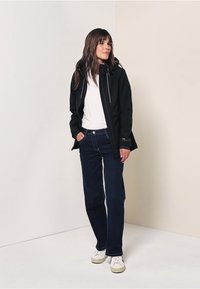 Zwarte waterdichte jas met rits aan de voorkant, gedragen over een wit shirt. Gecombineerd met donkere jeans en witte sneakers. Glad textuur, casual pasvorm.