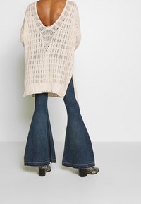 Pull en tricot beige clair avec un profond décolleté en V au dos, associé à un jean flare bleu foncé et des bottines à talons noires. La texture est en tricot ajouré.