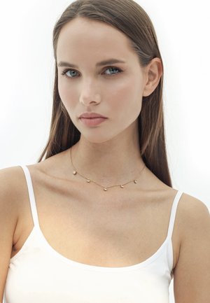 Goldene Halskette mit fünf kleinen, runden Perlen, die gleichmäßig auf einer zarten Kette verteilt sind, getragen über einem schlichten weißen Tanktop. Glatte Textur.