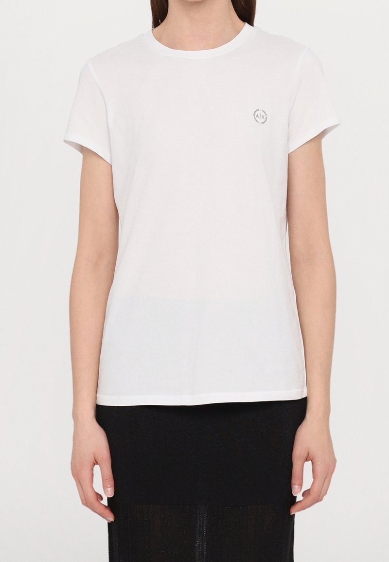 T-shirt en coton blanc avec un col rond, des manches courtes et un petit logo sur la poitrine gauche. Texture douce, coupe décontractée.