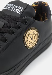 Baskets noires Versace Jeans Couture avec lacets noirs, patch logo beige rond et doublure intérieure à motifs.