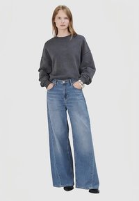 Übergroßes dunkelgraues Sweatshirt mit überschnittenen Schultern, kombiniert mit weit geschnittenen hellblauen Jeans. Das Model steht mit den Händen in den Taschen vor einem schlichten Hintergrund.