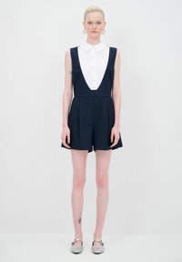Monosuit sin mangas de color azul marino con un cuello blanco de botones, que presenta un escote en V profundo, una cintura estructurada y pantalones cortos amplios. Calzado plateado que acentúa.