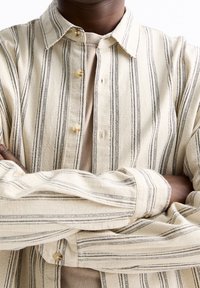 Chemise beige à rayures boutonnée en tissu léger, avec des rayures noires verticales, un col et des boutons sur le devant et les poignets.