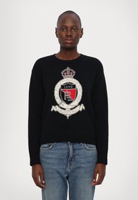 Zwarte gebreide trui met een geborduurd wapenschild met een kroon, rode en witte accenten, en een ribgebreide zoom. Gecombineerd met blauwe spijkerbroeken.