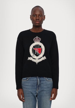Maglione nero a maglia con un ricamo che presenta uno stemma con una corona, dettagli rosso e bianco, e orlo a coste. Abbinato a jeans in denim blu.