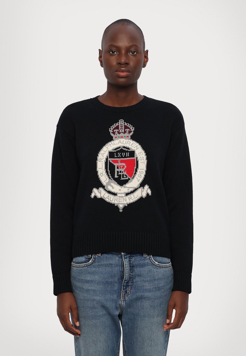 Zwarte gebreide trui met een geborduurd wapenschild met een kroon, rode en witte accenten, en een ribgebreide zoom. Gecombineerd met blauwe spijkerbroeken.