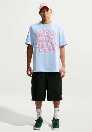 Uomo che indossa una maglietta azzurro chiaro con motivo circolare rosa, pantaloncini neri, calzini bianchi Nike, scarpe da ginnastica verdi e un cappellino rosa chiaro, in piedi davanti a uno sfondo semplice.