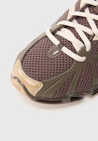 Lähikuva ruskeasta ja beigestä urheilukengästä, jossa on verkkopäällinen, kermaiset nauhat ja vahvistettu kärki valkoisella taustalla.