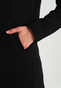 Vestido negro texturizado con un patrón acolchado, que presenta mangas largas y un bolsillo lateral, mostrando una silueta ajustada.