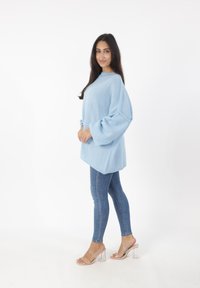 Helles blaues, übergroßes Strickoberteil mit weiten Puffärmeln, kombiniert mit enganliegenden Denim-Jeans und klaren Absatzsandalen, vor einem schlichten Hintergrund.