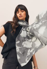 Harmaa kukkakuvioinen huivi, jossa on teksturoitu, ribbineulosta muistuttava kuvio sileää taustaa vasten; yhdistetty mustaan neulottuun yläosaan ja tummiin housuihin.