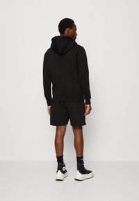 Sudadera negra con un bolsillo canguro, combinada con pantalones cortos negros. La persona lleva zapatillas altas negras y calcetines negros.