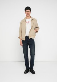 Bézs könnyű bomber dzseki fehér, piros feliratos pólóval, sötét farmernadrággal és fekete cipővel, egyszínű háttér előtt.