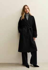 Gabardina negra con diseño de doble botonadura, cintura con cinturón, solapas anchas y bolsillos laterales, combinada con un suéter de cuello alto gris y pantalones anchos oscuros.