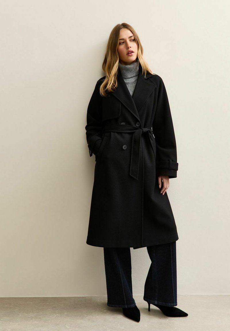 Gabardina negra con diseño de doble botonadura, cintura con cinturón, solapas anchas y bolsillos laterales, combinada con un suéter de cuello alto gris y pantalones anchos oscuros.