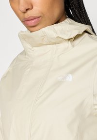Veste imperméable beige clair avec un col montant, présentant un logo blanc sur le côté gauche de la poitrine et un tissu lisse et texturé.