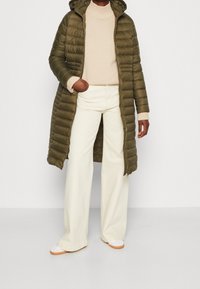 Persoon in een olijfgroene lange pufferjas over een Beigebruine trui, crèmekleurige wijde broek en witte sneakers met gumrubberen zolen.