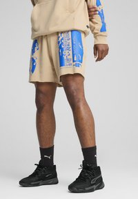Beige shorts med blå grafiske designs, ufuldstændig kant og en løs pasform. Kombineret med sorte sneakers og mørke sokker.