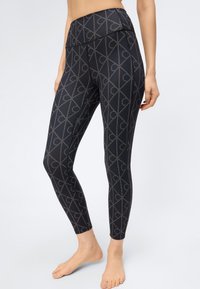 Γυναίκα που φοράει μαύρο κολάν ψηλής μέσης με γεωμετρικό σχέδιο από διασταυρούμενες γραμμές και σχήματα σεληνοειδή, όρθια ξυπόλυτη πάνω σε λευκό φόντο.