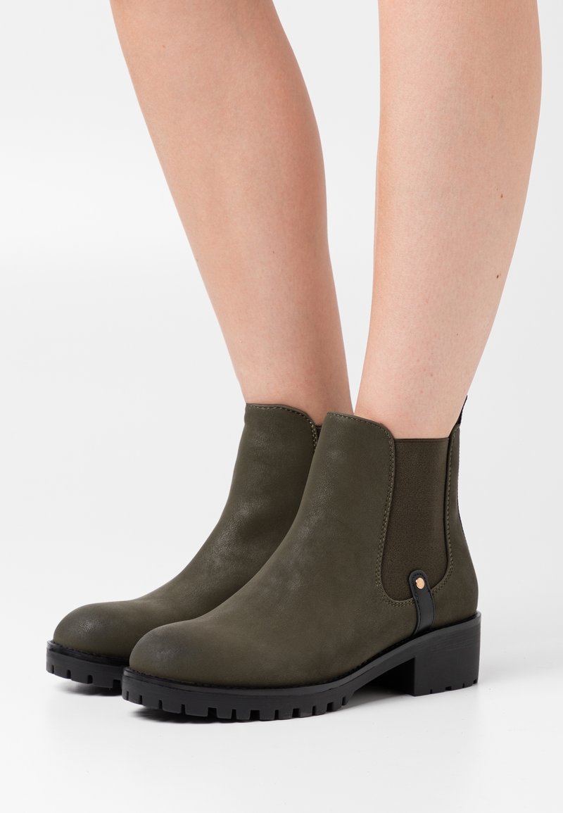 Bottes montantes vert olive mat avec des panneaux latéraux élastiques noirs et des semelles épaisses à crampons, portées sur des jambes nues.