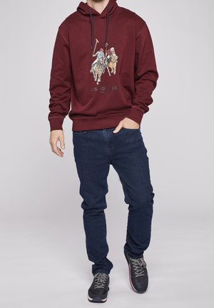 Homme portant un sweat à capuche bordeaux avec un motif joueur de polo, jean bleu foncé et baskets noires, debout avec une main dans la poche.