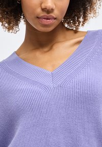 Közeli kép egy személyről, aki texturált levendulalila V-nyakú pulóvert visel, látható a nyaka, a kulcscsontja és a göndör haja egy része.