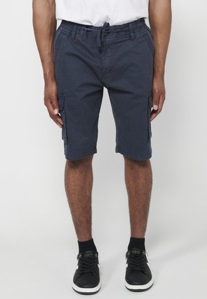 Mann trägt marineblaue Cargo-Shorts mit elastischem Bund und Kordelzug, schwarze Socken und schwarze Sneakers mit weißen Sohlen.