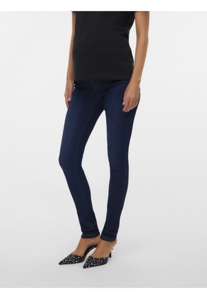 Personne portant un jean skinny bleu foncé, un haut noir et des escarpins noirs à bouts pointus avec des pois blancs, debout devant un fond blanc.