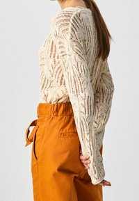 Beige neulepusero, jossa on pitsikuvio ja pitkät hihat, yhdistettynä oransseihin korkeavyötäröisiin housuihin, joissa on nauhavyö ja sivutaskut.