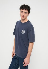 Tamsiai mėlynas medvilninis marškinėlis trumpomis rankovėmis ir apvalia iškirpte, su baltu grafiniu atspaudu kairėje krūtinės pusėje.