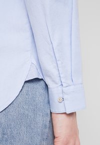 Hellsblaues Hemd aus strukturiertem Stoff, mit leicht abgerundetem Saum und einem knöpfbaren Bund. Vor einem Denim-Hintergrund positioniert.