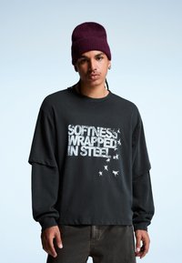 Fekete, oversized hosszú ujjú póló, textúrált részletekkel és fehér grafikai nyomattal: "PUHASÁG ACÉLBAN CSOMAGOLVA", csillagok kíséretében.