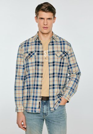 Młody mężczyzna w niezamkniętej koszuli w niebiesko-beżowo-białą kratę, nałożonej na beżowy t-shirt z nadrukiem, oraz w jasnoniebieskich dżinsach, z jedną ręką w kieszeni.