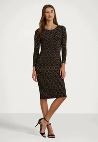 Γυναίκα που φοράει ένα μαύρο, φαρδύ, μακρυμάνικο φόρεμα μέχρι το γόνατο με χρυσό κυκλικό σχέδιο, φορώντας μαύρα ψηλοτάκουνα παπούτσια με λουράκι στον αστράγαλο, που στέκεται σε έναν λευκό φόντο.