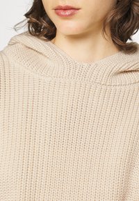 Felpa con cappuccio beige a maglia con una trama testurizzata, caratterizzata da un'ampia scollatura e un aspetto morbido e accogliente. Orlo leggermente rialzato e dettagli del cappuccio.