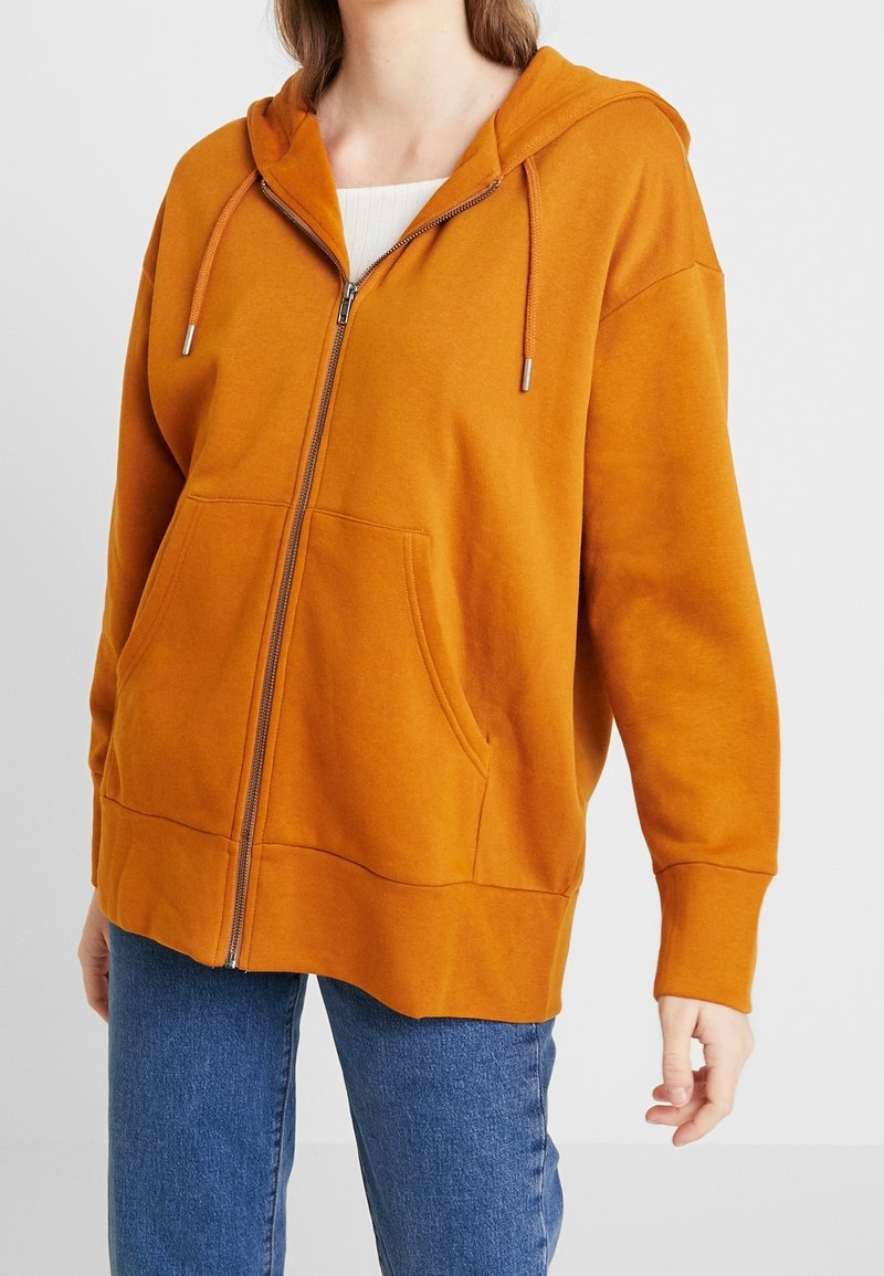 Sudadera con capucha naranja y cierre de cremallera, con dos bolsillos frontales, puños acanalados y un acabado texturizado, combinada con vaqueros azules.