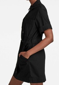 Kvinna som bär en svart skjortklänning med kort ärm, krage, framfickor och dragsko i midjan, med handen vilande i en ficka.