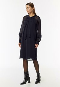 Marineblaues Kleid mit transparenten Ärmeln, einem gebundenen Kragen und einer lässigen Passform, kombiniert mit schwarzen Stiefeletten und transparenten dunklen Strumpfhosen.