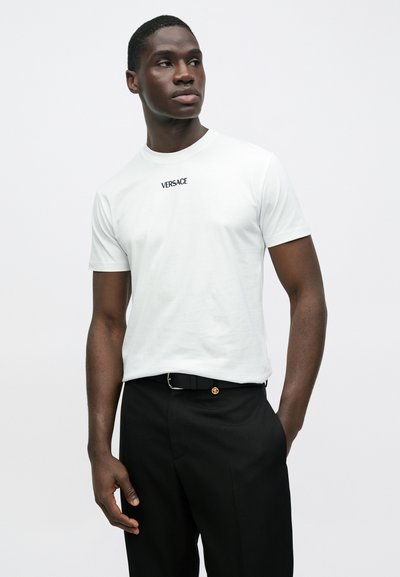 Biała bawełniana koszulka z czarnym logo „VERSACE” na piersi, zestawiona z czarnymi spodniami i czarnym paskiem z złotą, okrągłą klamrą.