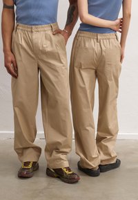 Beige, loose-fitting pants with an elastic waistband, pleats at the front, and back pockets; worn with sneakers; cotton material.
