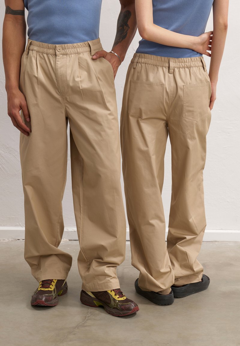 Beige, loose-fitting pants with an elastic waistband, pleats at the front, and back pockets; worn with sneakers; cotton material.