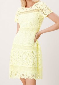 Nainen, joka on pukeutunut lyhythihaiseen, polvipituiseen vaaleankeltaiseen pitsimekkoon, jossa on kukkakuoseja ja verkkoyksityiskohtia, poseeraa käsi lanteellaan.