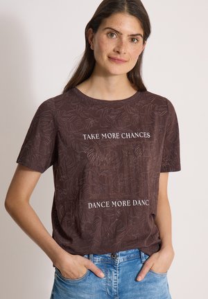 Kvinde med langt brunt hår bærer en mørk mønstret T-shirt med teksten "TAKE MORE CHANCES, DANCE MORE DANCES," og blå jeans, med hænderne i lommerne.