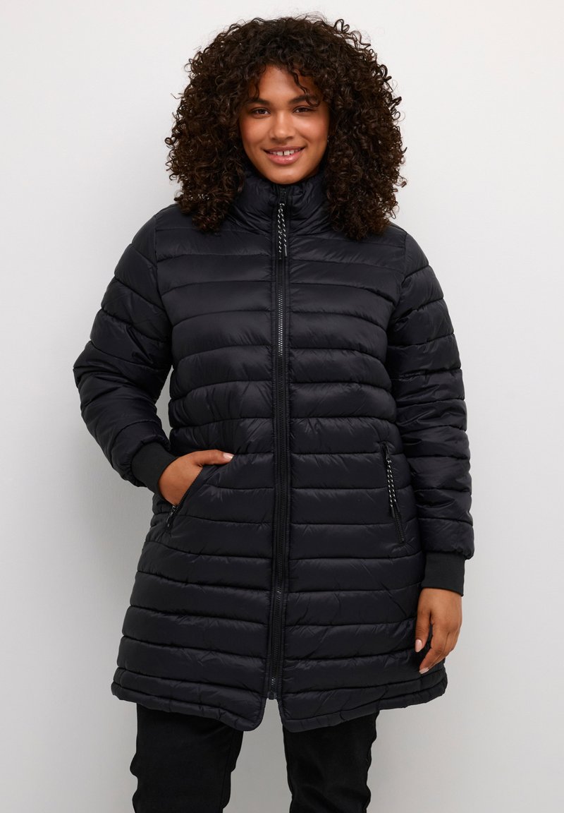 Lächelnde Frau mit lockigem Haar, die eine schwarze, wattierte Langsteppjacke mit Reißverschlusstaschen trägt, vor einem einfarbigen, hellen Hintergrund stehend.