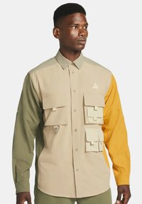 Tvåttonad skjorta med beige, olivgrön och senapsgul sektioner; har flera framfickor och en logotyp på bröstet.