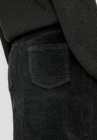 Gros plan d'une personne portant un pantalon en velours côtelé noir avec une poche arrière et un pull en maille texturé foncé.