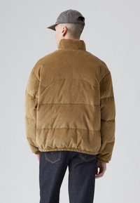 Brun corduroy pufferjakke med høj krave, elastisk kant og quiltet design. Båret med mørke denimjeans, vist fra ryggen.